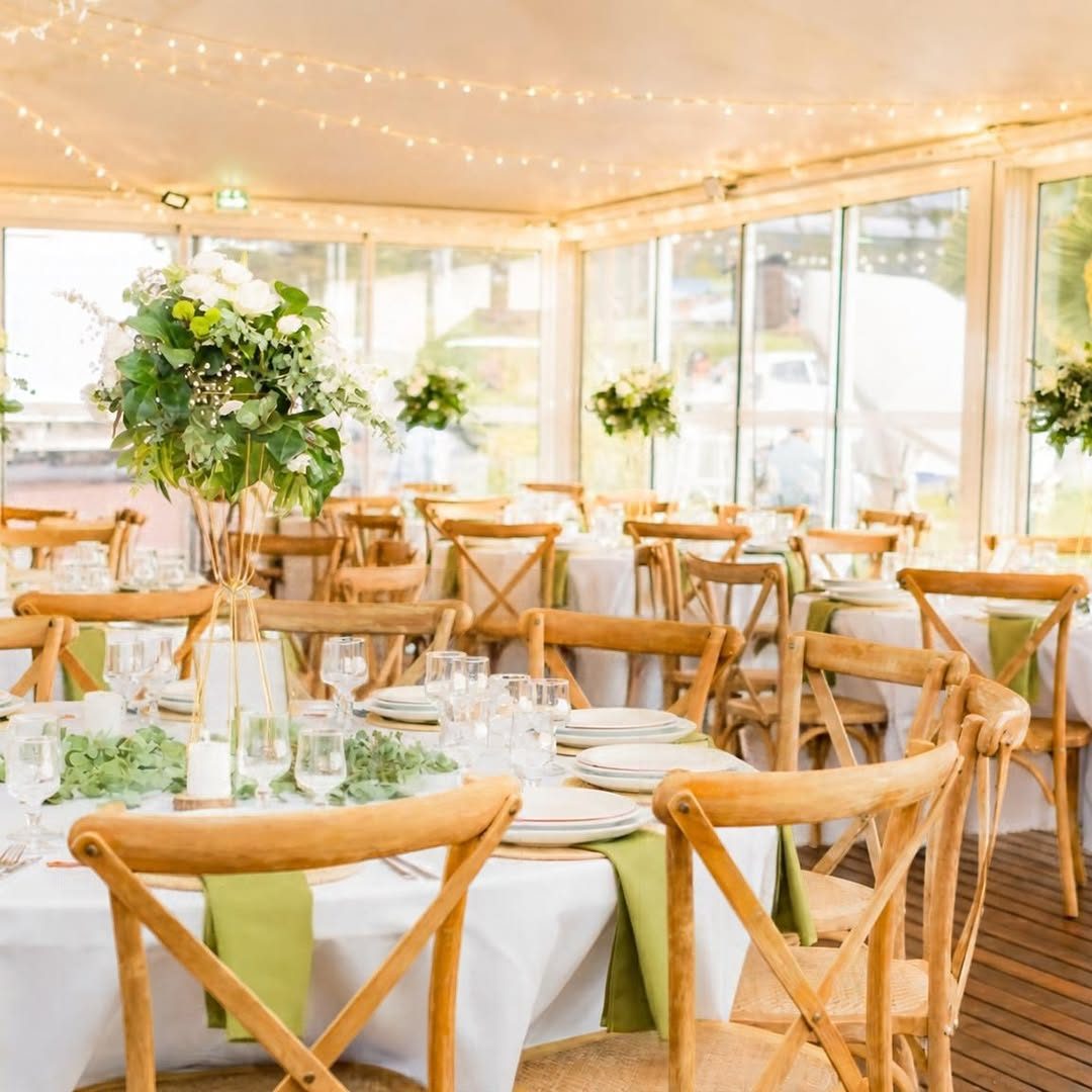 Salle de réception décorée : tables rondes nappées de blanc, chaises en bois croisées, compositions florales vertes et guirlandes lumineuses